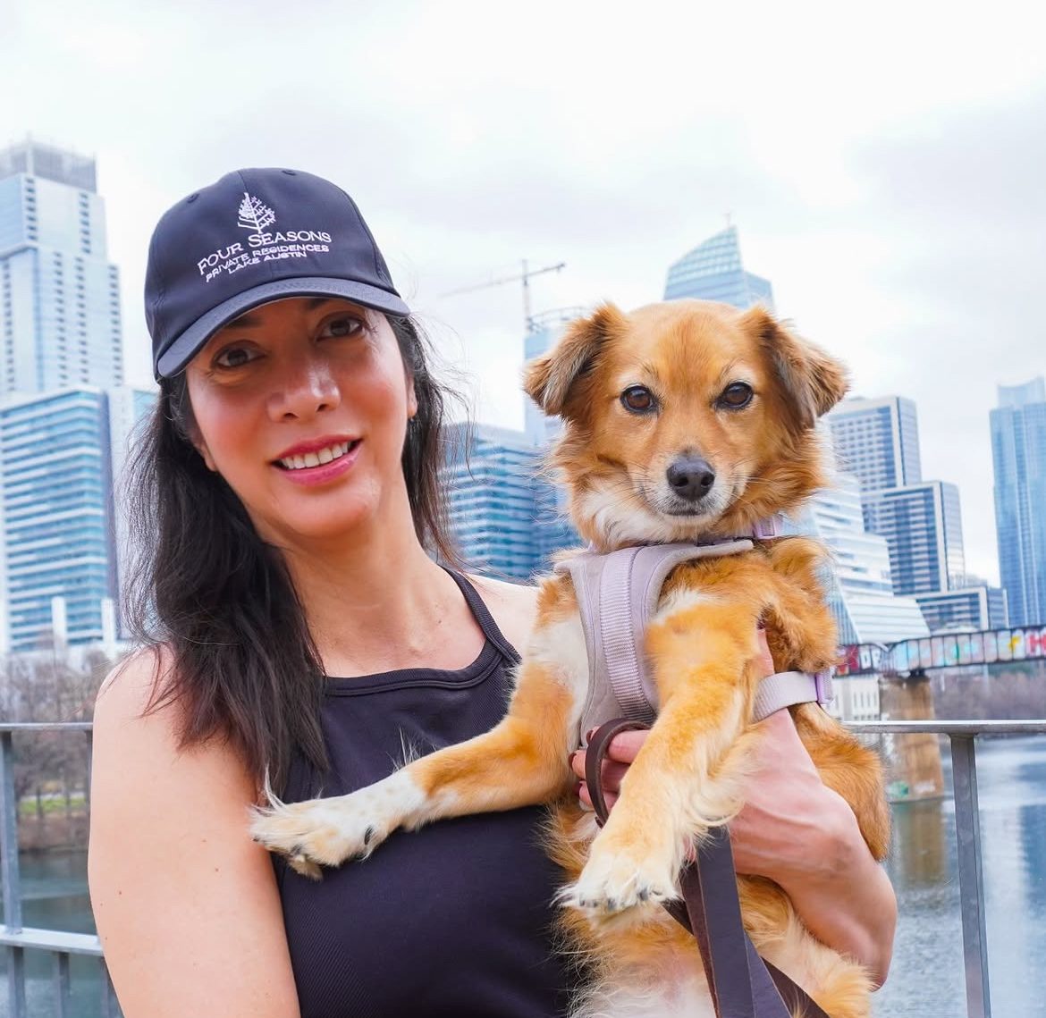 Liz Reyna holding Lady, a small rescue dog, on the Lady Bird Lake trail with the downtown Austin skyline behind them